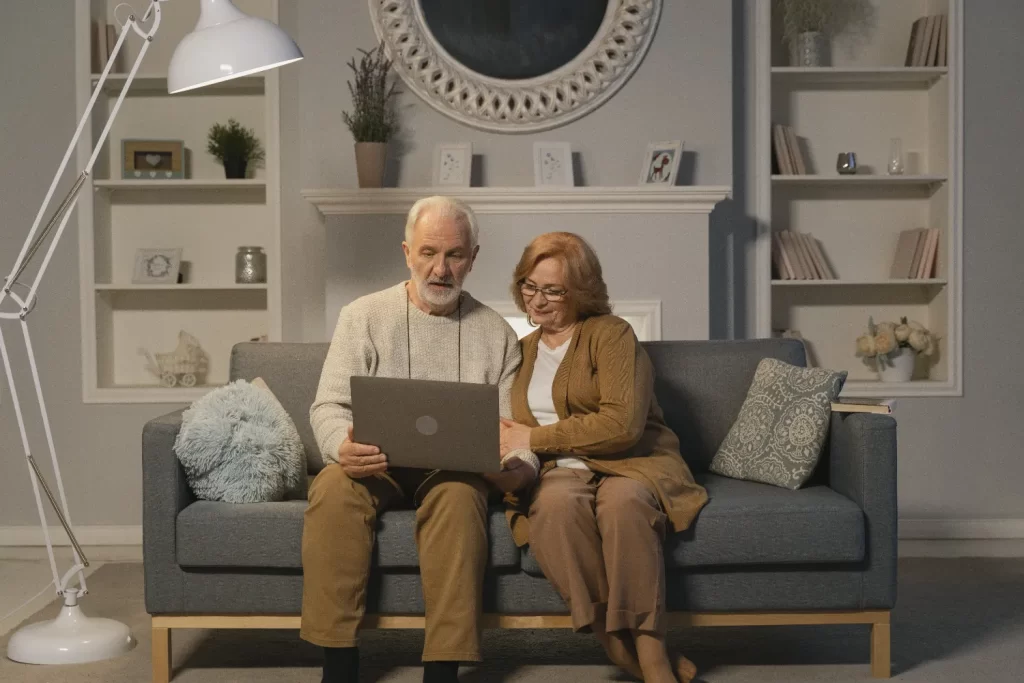 زوج سالمند که بر روی مبل نشسته اند و مشغول به لپ تاپ هستند و در پشت آن ها ساعت و دکور چوبی که در آن کتاب قرار دارد وجود دارد
