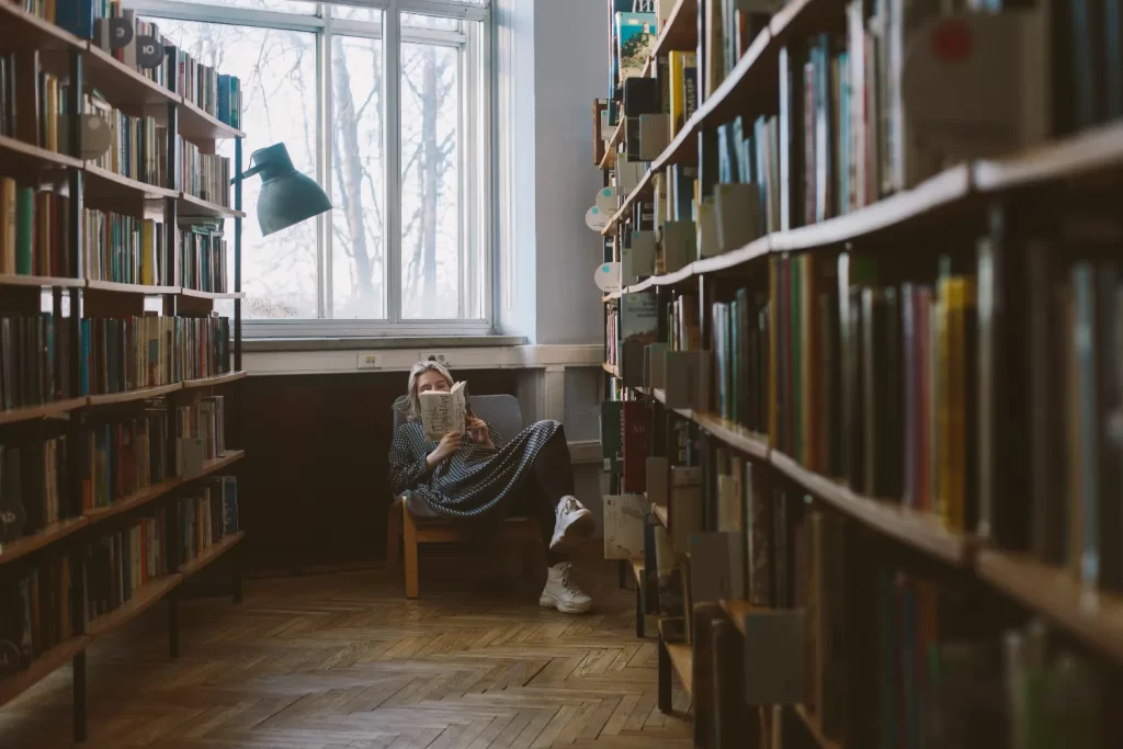 فردی که در کتابخانه بر روی مبل نشسته است و در حال خواندن کتاب میباشد و در پشت آن پنجره باز شده قرار دارد و د سمت چپ و راست آن بخش کتاب ها قرار دارد