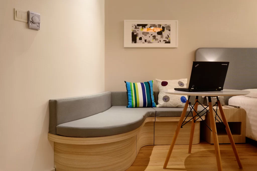 مبل چوبی راحتی که در کنار آن تخت خواب و در روبه روی آن لپ تاپ بر روی میز قرار دارد