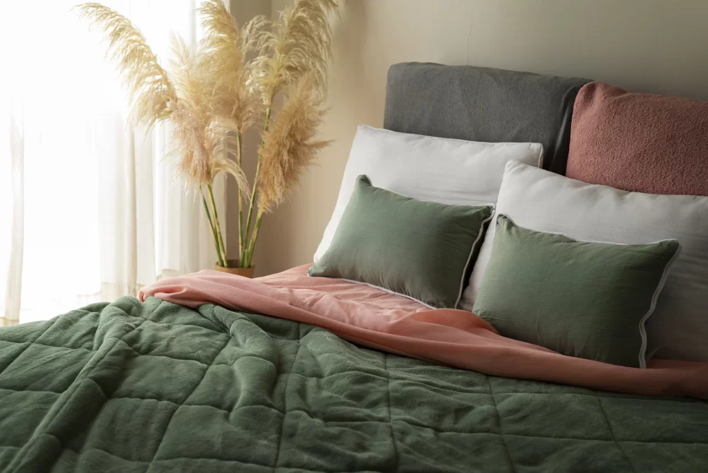 تخت خواب با پتو سبز و بالشت های سبز و سفید که در کنار آن گل و پرده قرار دارد