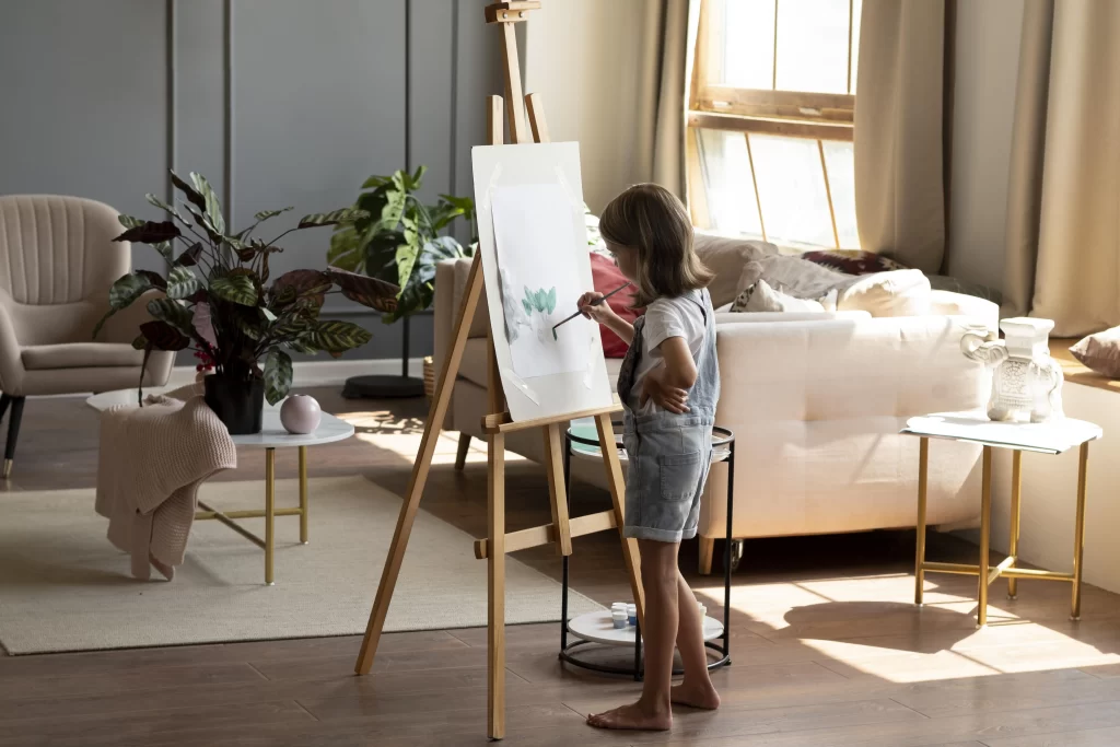 دختر بچه ای که در هال قرار دارد و درحال نقاشی کشیدن میباشد و کاغذ را بر روی پایه نقاشی گذاشته است و در پشت پایه مبل های راحتی کرمی و طوسی و میز عسلی که بر روی آن گل قرار دارد وجود دارد