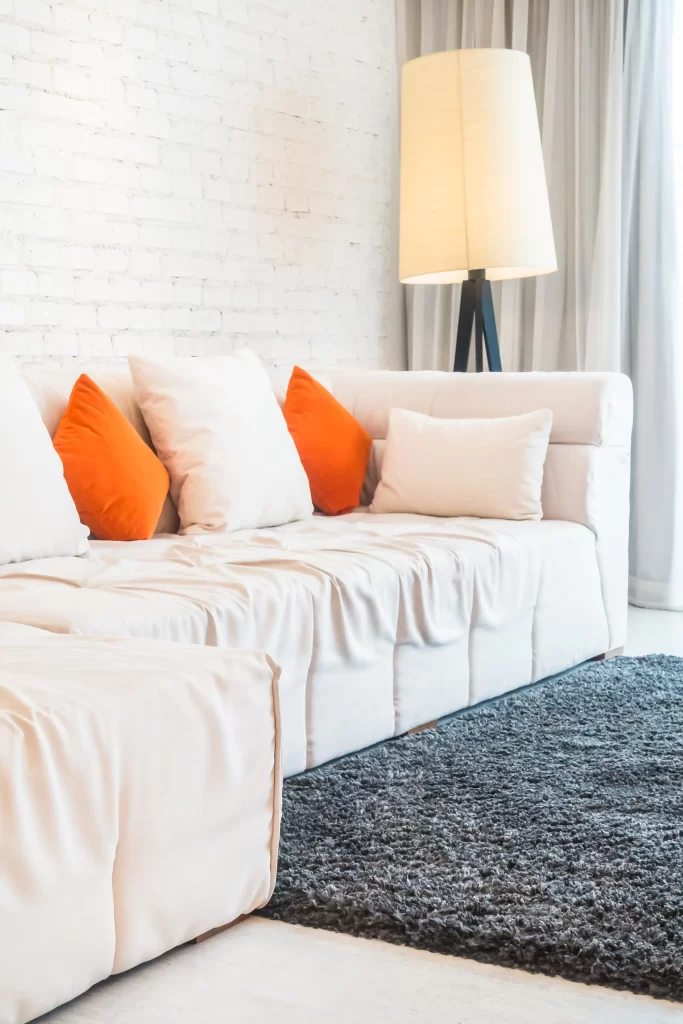 مبل راحتی و تخت شو سفید که بالشت های نارنجی و سفید بر روی آن قرار دارد و در کنار مبل نیز آباژور قرار دارد